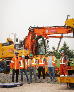 6 male operating engineers stand side by side smiling at the camera in front of a High Rail Excavator.