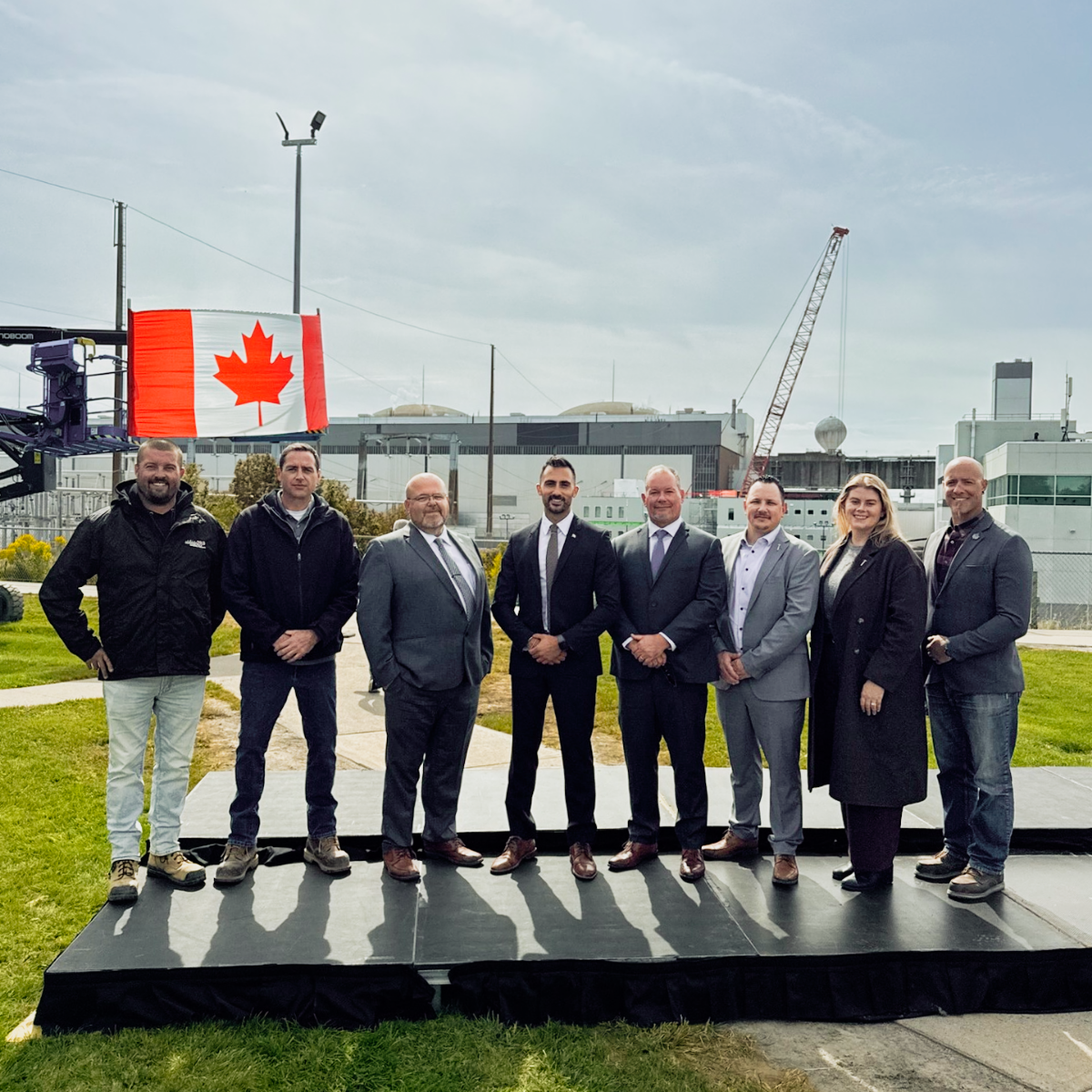 A group of men and 1 women stand side by side with the Canadian Flag behind them