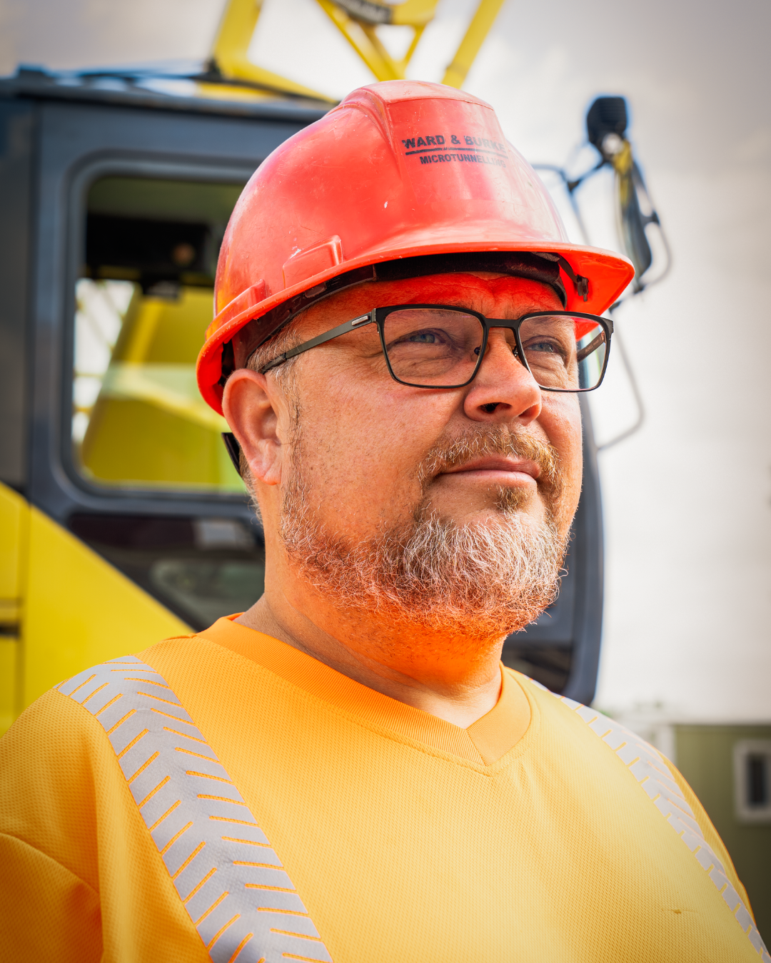 A close up picture of a man staring off into the distance, he wears an orange P.P.E shirt and red hard hat