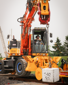 A male operating engineer sits inside a High Rail Excavator facing the camera, the machine lifts a 240lbs concrete block