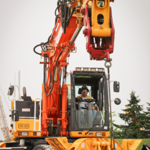 A male operating engineer sits inside a High Rail Excavator facing the camera, the machine lifts a 240lbs concrete block