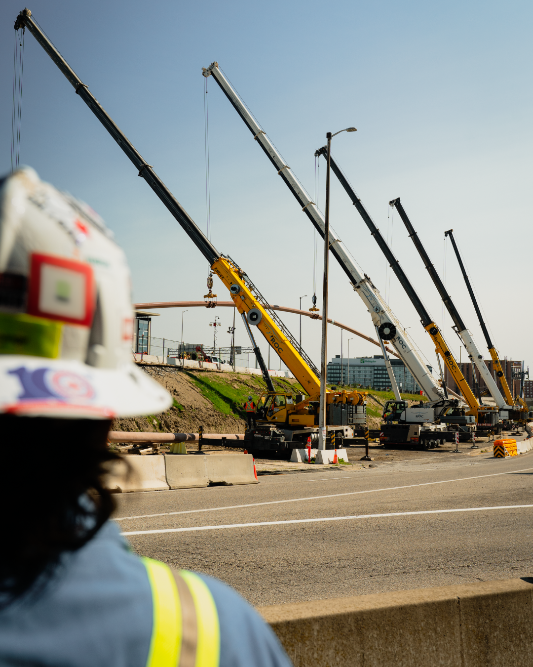 5 cranes as the focus of the photo with a semi-blurry picture of the back of a man wearing a white hard hat's head in the foreground
