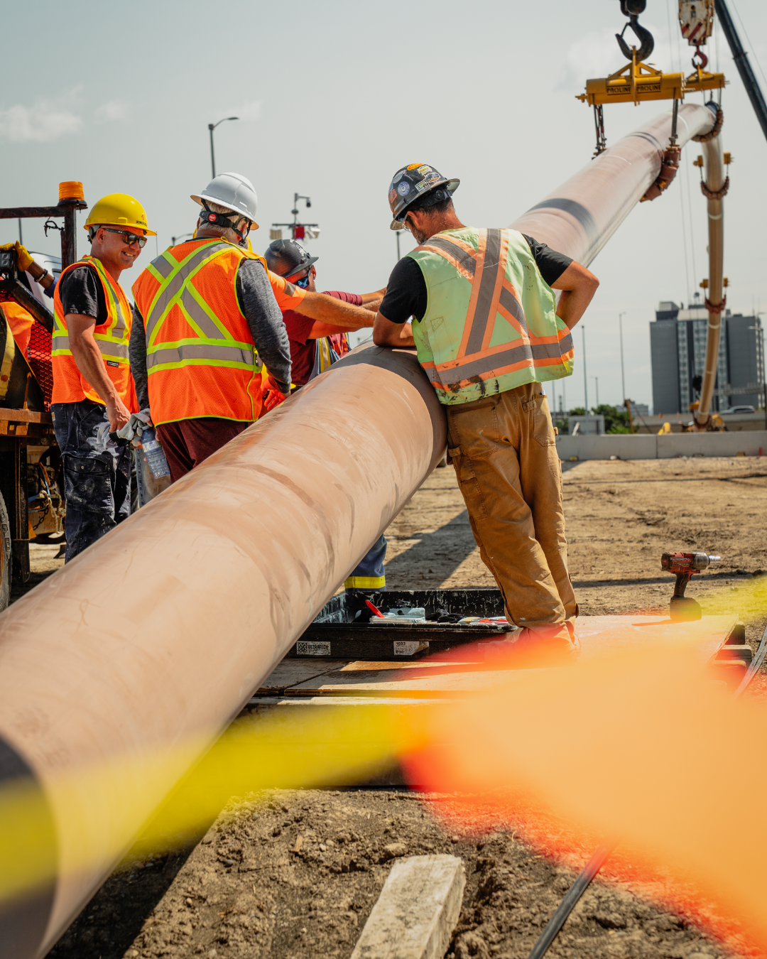 4 construction workers looking over a pipe being held just above the ground by a crane