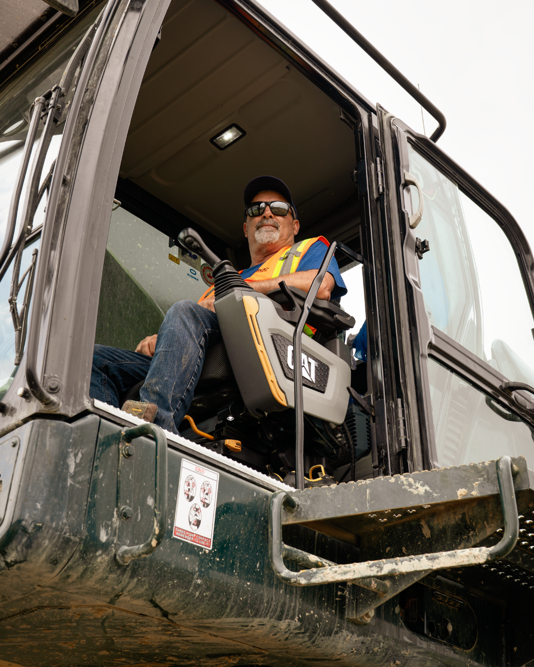 A man sits inside of an excavator looking downwards at the camera.