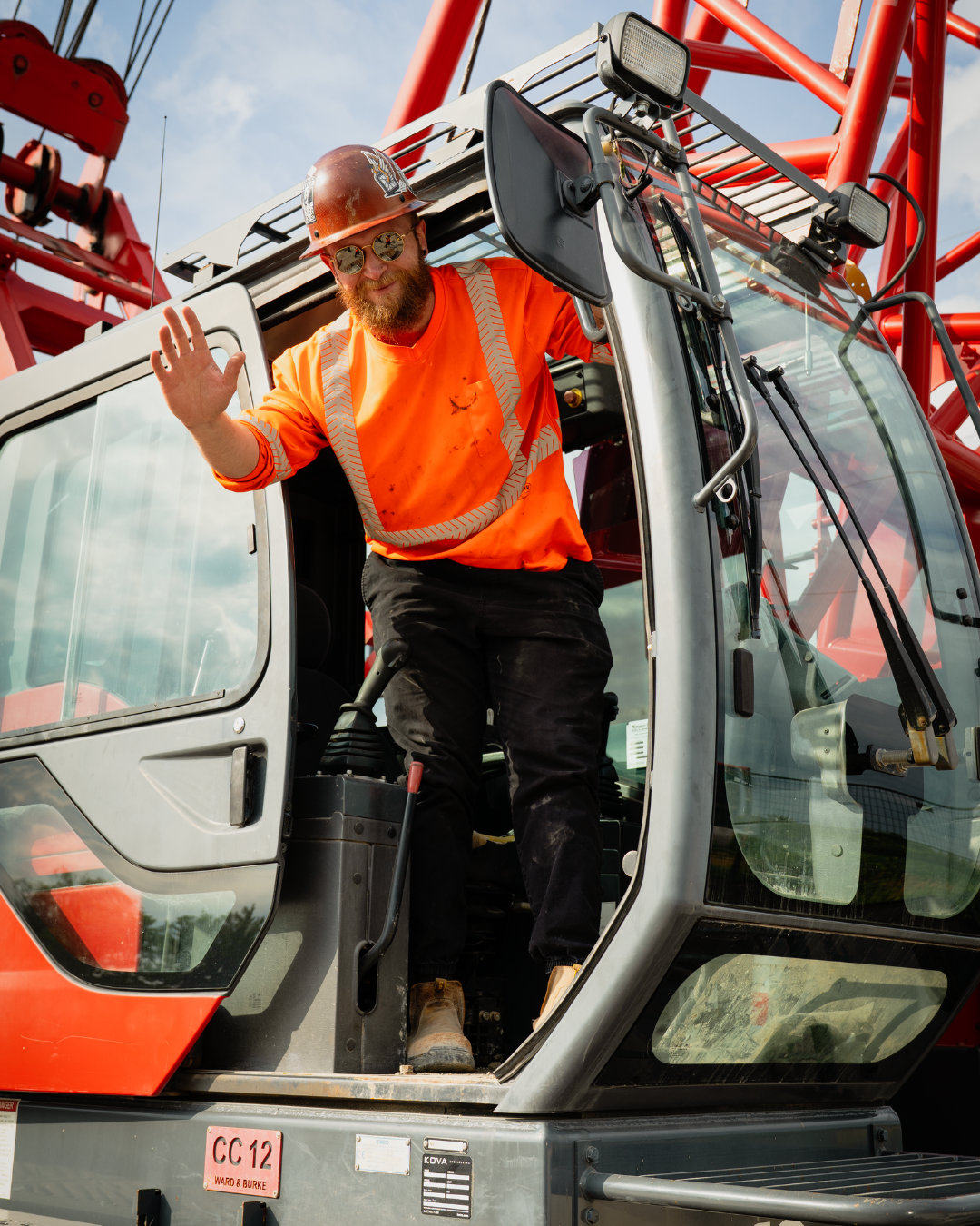 A construction worker is stepping out of a crane wearing a red hard hat and an orange P.P.E shirt, waving at the camera.