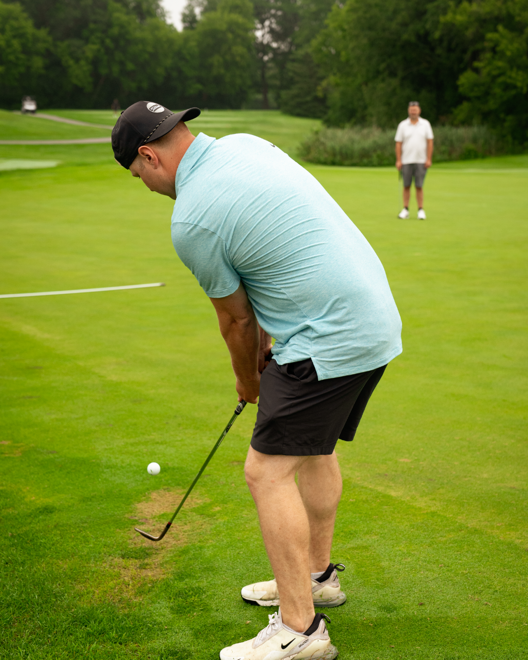 A man in a blue t-shirt has his back turned to the camera and is preparing to hit a golf ball.
