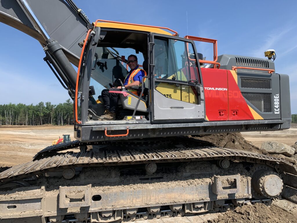 Member sitting inside operating the excavator, pictured from the side view.
