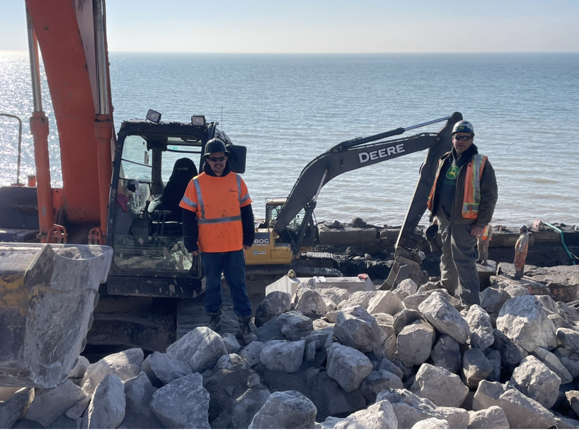 Two members standing in front of excavator on the shoreline
